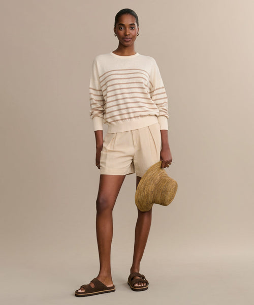 A woman stands against a beige background wearing the Avery Crewneck, a lightweight knit striped sweater, paired with beige shorts, brown sandals, and holding a woven sun hat.