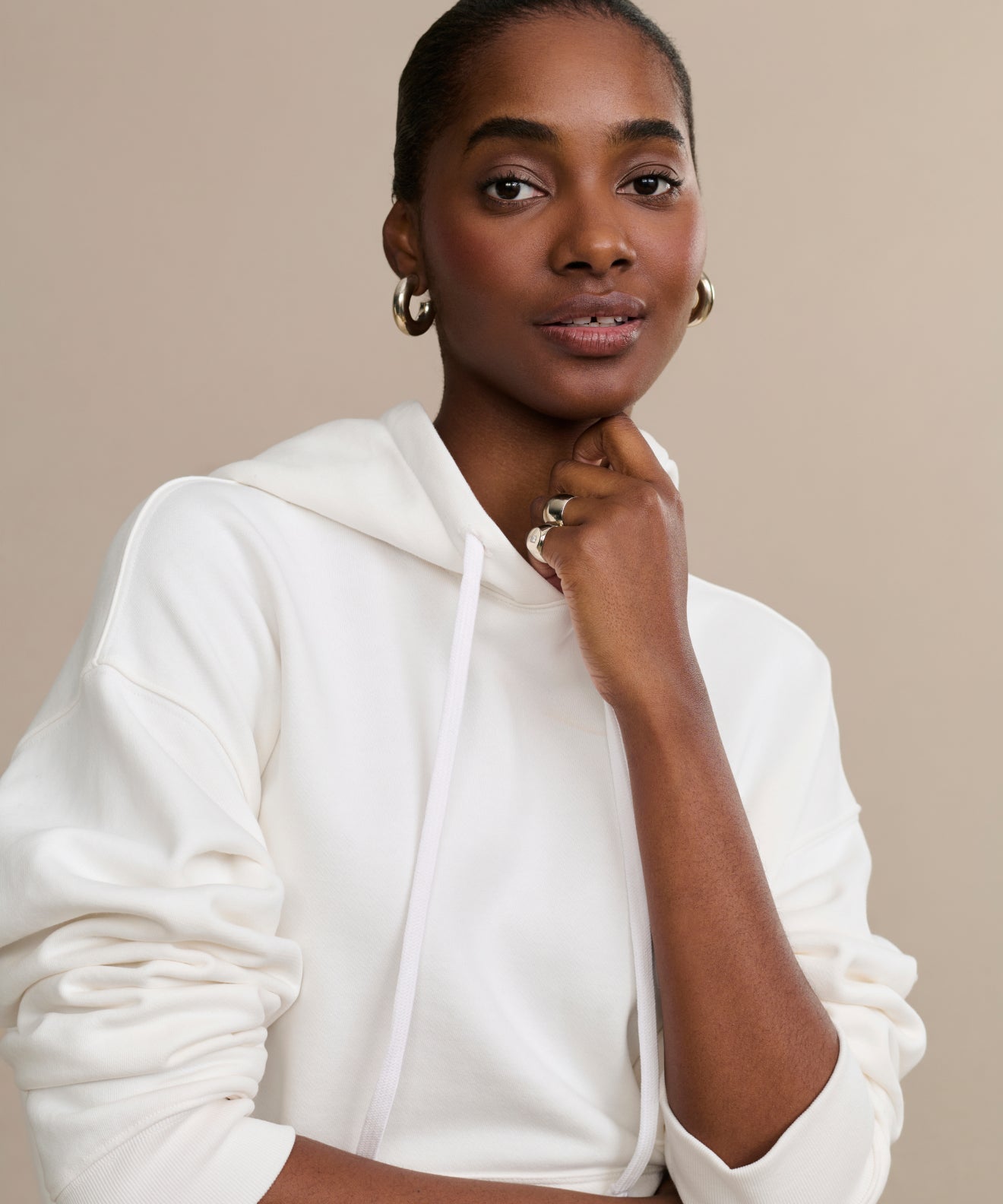 Dressed in the Bay Hoodie and gold hoop earrings, a person poses confidently with one hand under their chin, gazing at the camera against a plain beige background.