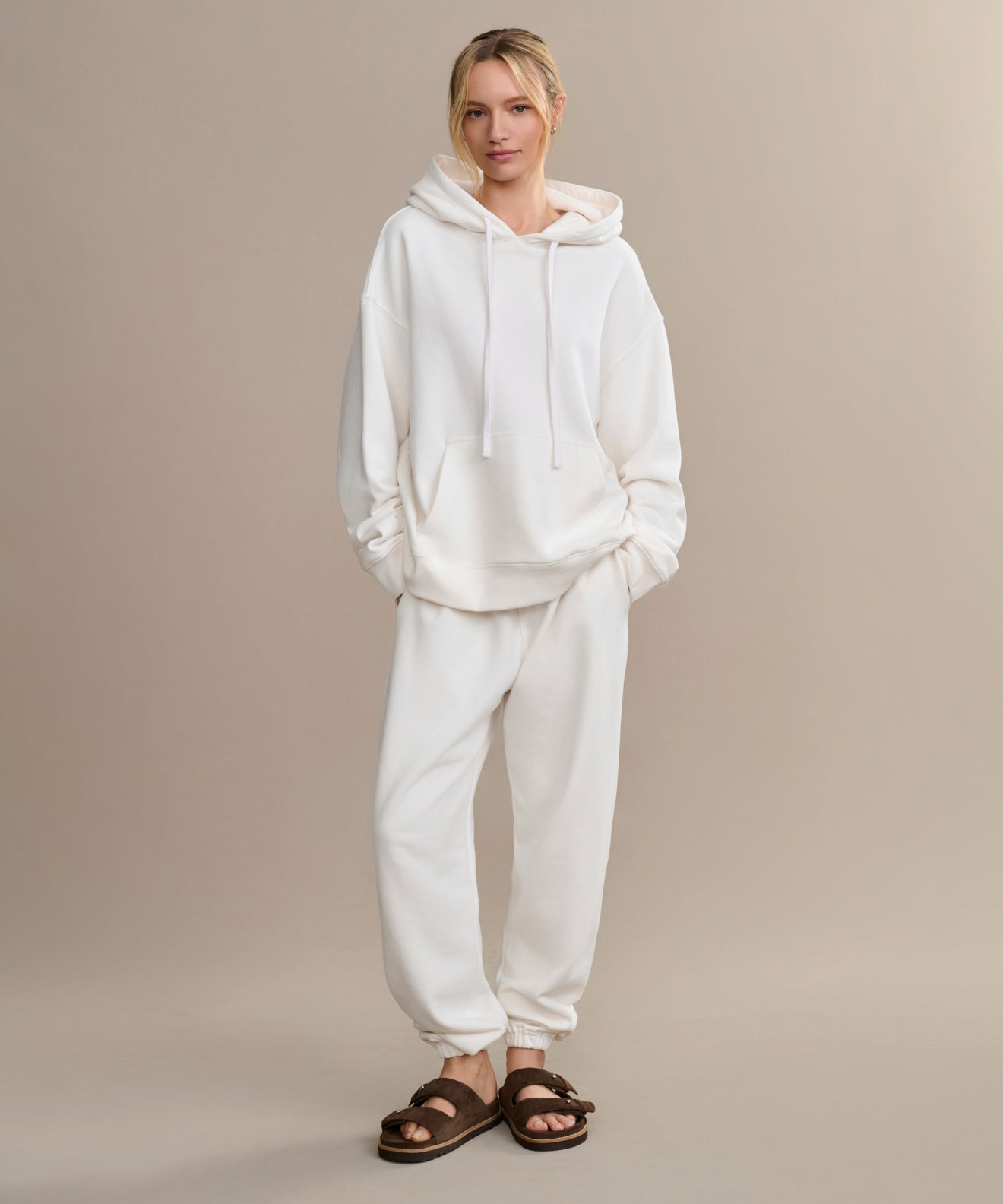 A person stands against a beige background wearing an oversized Bay Hoodie in ivory, matching white sweatpants, and brown fuzzy sandals—perfect for a cozy wardrobe.