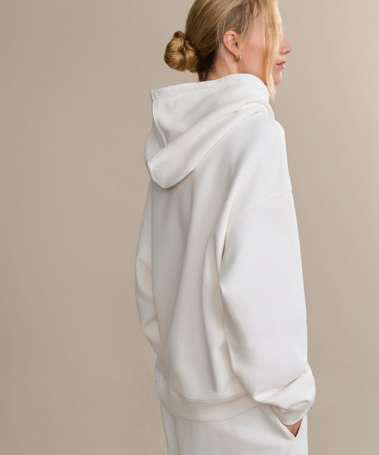 A woman with blonde hair in a bun wears the ivory Bay Hoodie and matching pants, standing in profile with her hands in her pockets—a cozy wardrobe essential—against a neutral beige background.