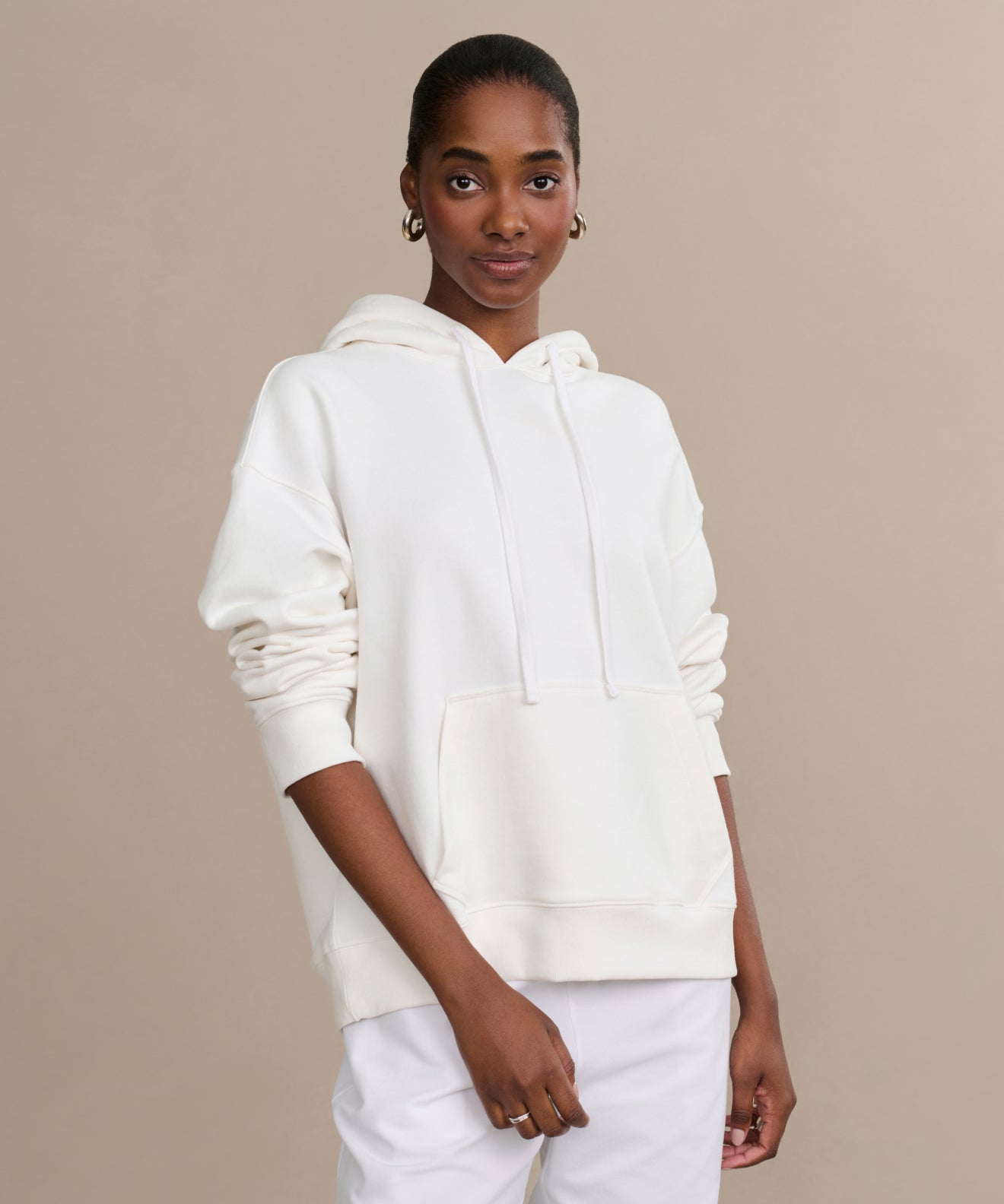 A woman stands against a beige background wearing the Bay Hoodie in ivory and white pants, looking at the camera with a slight smile and her hands relaxed by her sides—a perfect cozy wardrobe addition.