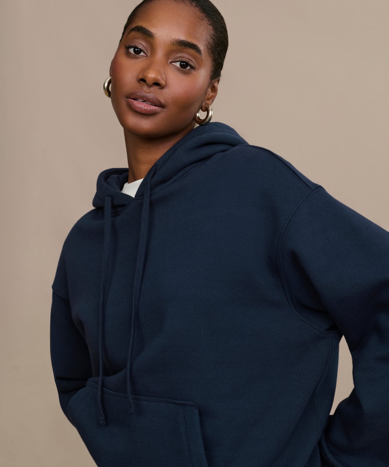 Wearing the Bay Hoodie, a woman with silver hoop earrings stands confidently against a beige background. The brushed fleece fabric of the hoodie adds comfort to her casual and stylish look.