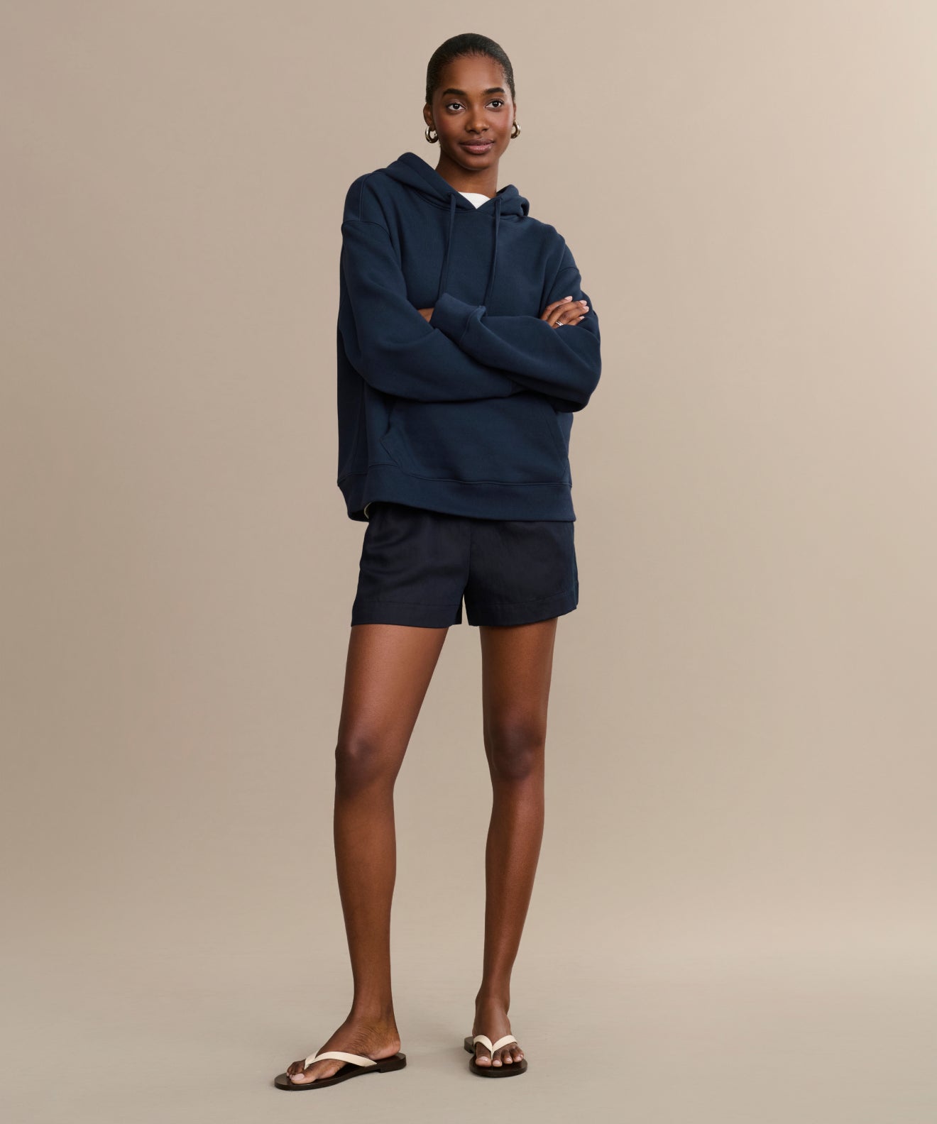 A woman smiles softly at the camera, arms crossed, wearing a Navy Bay Hoodie and black shorts with white sandals against a plain beige background.