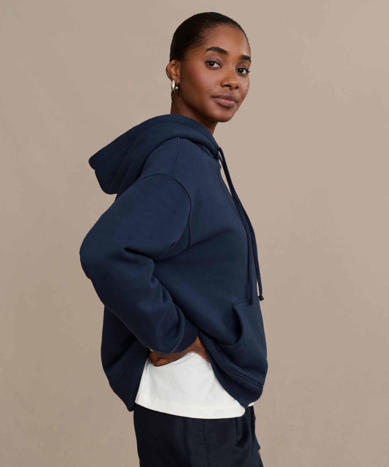 A woman with her hair pulled back poses against a beige background, looking at the camera. She wears a navy Bay Hoodie over a white shirt.