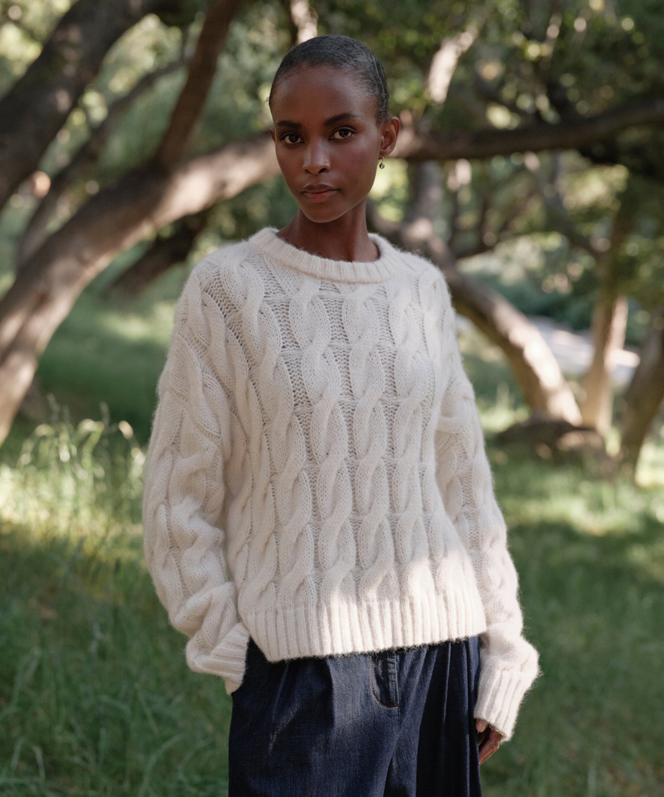 A woman with short hair wears the Cable Serena Crewneck in ivory with dark pants, standing outdoors in a sunlit wooded area surrounded by green grass and trees.