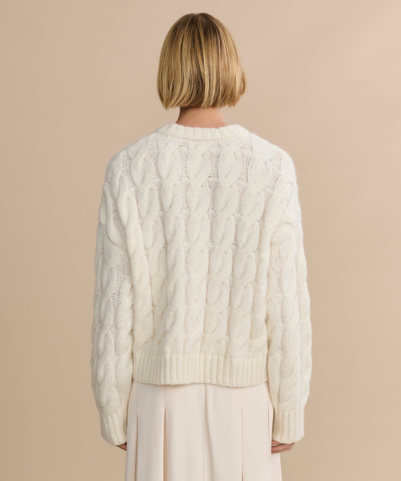 A person with short blonde hair is seen from behind, wearing the Cable Serena Crewneck in ivory and a light pleated skirt, standing against a beige background.
