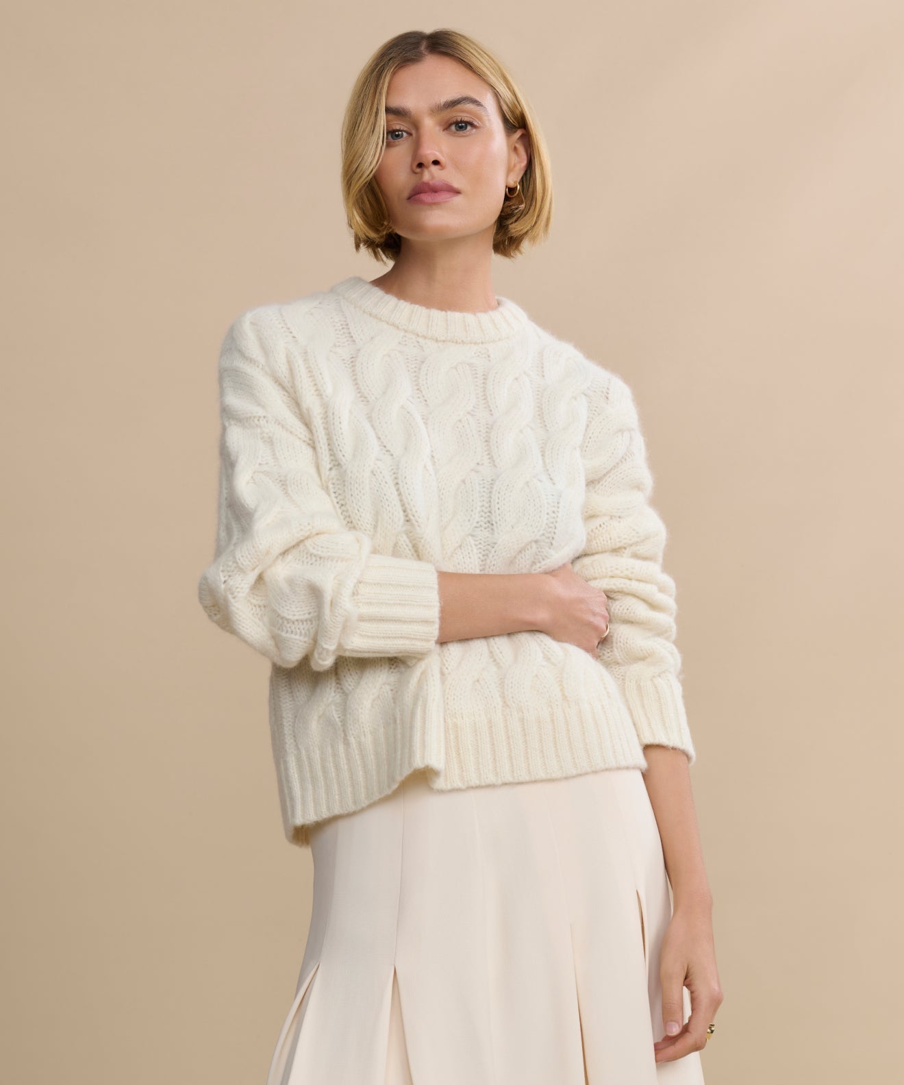 A woman with short blonde hair wears the Cable Serena Crewneck in ivory and a light pleated skirt, standing against a beige background with a neutral expression.