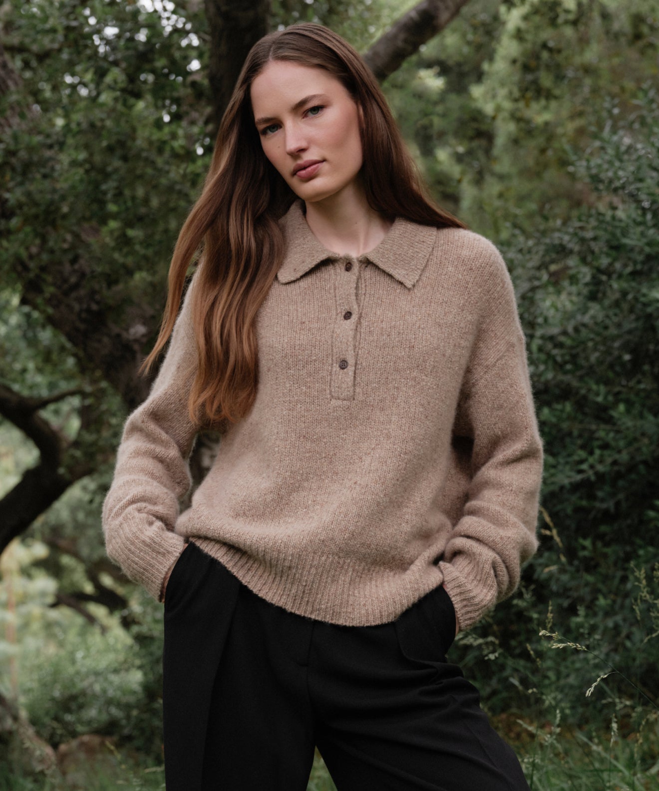 A woman with long brown hair stands outdoors among green trees, wearing the Cashmere Burke Polo and black pants, her hands in her pockets and a calm expression on her face.