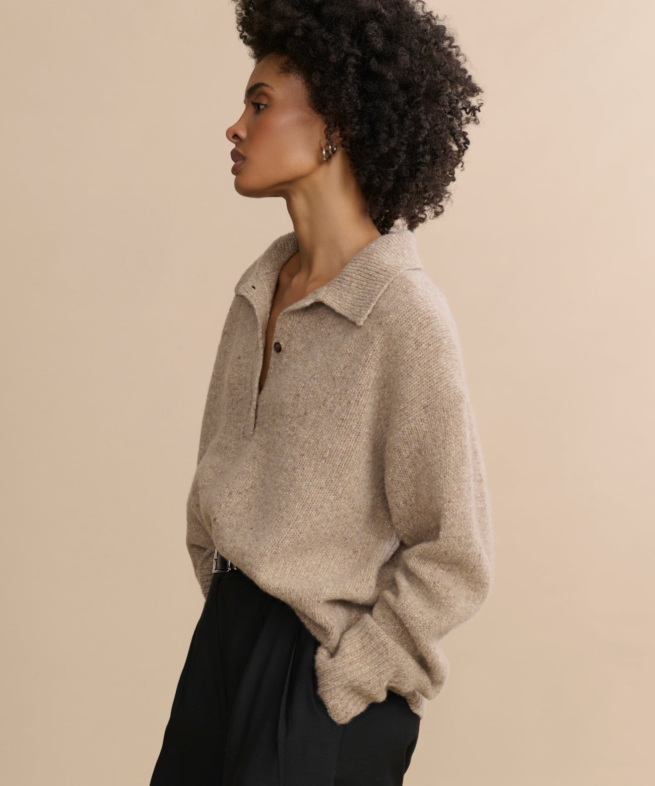 A woman with short curly hair poses in profile against a beige background, wearing the oversized Cashmere Burke Polo sweater with buttons and black pants, her hands casually tucked in her pockets.