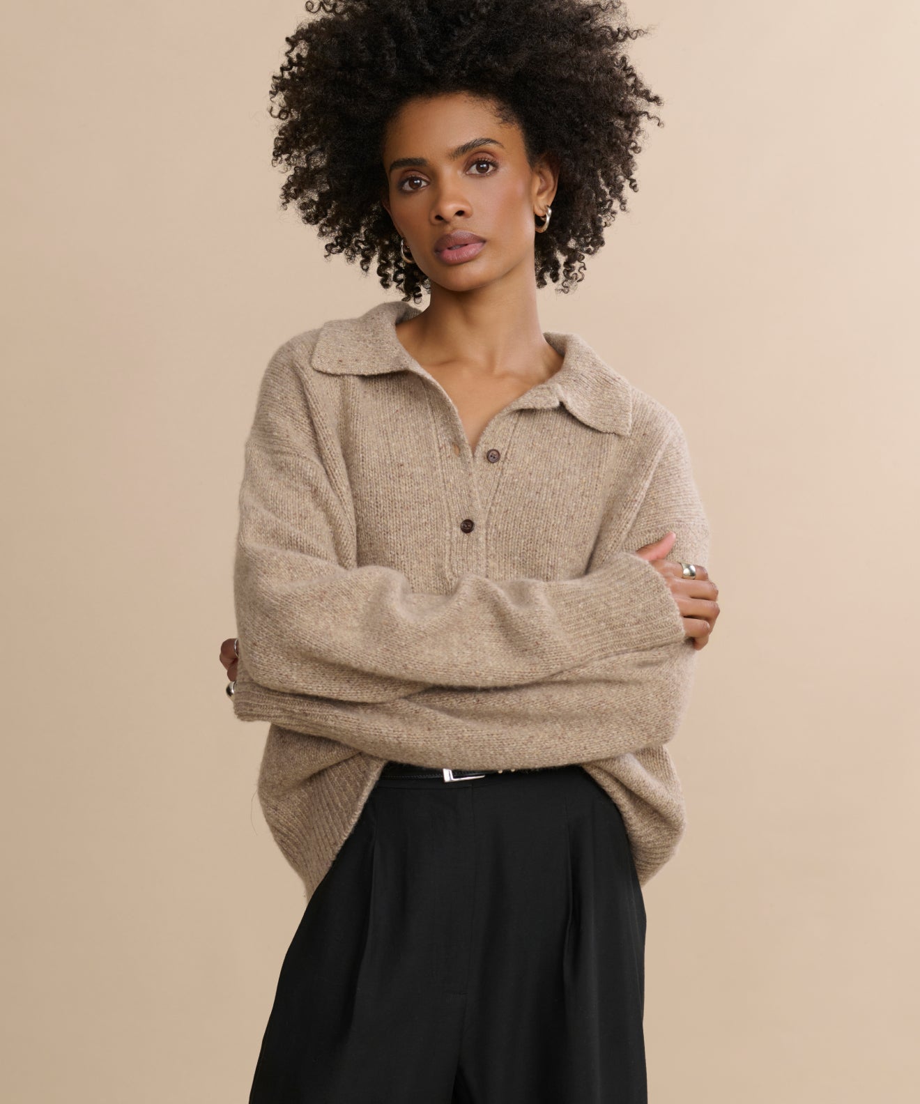 A person with curly hair wears the Cashmere Burke Polo, styled oversized with large sleeves and black pants, standing with arms crossed against a plain beige background.