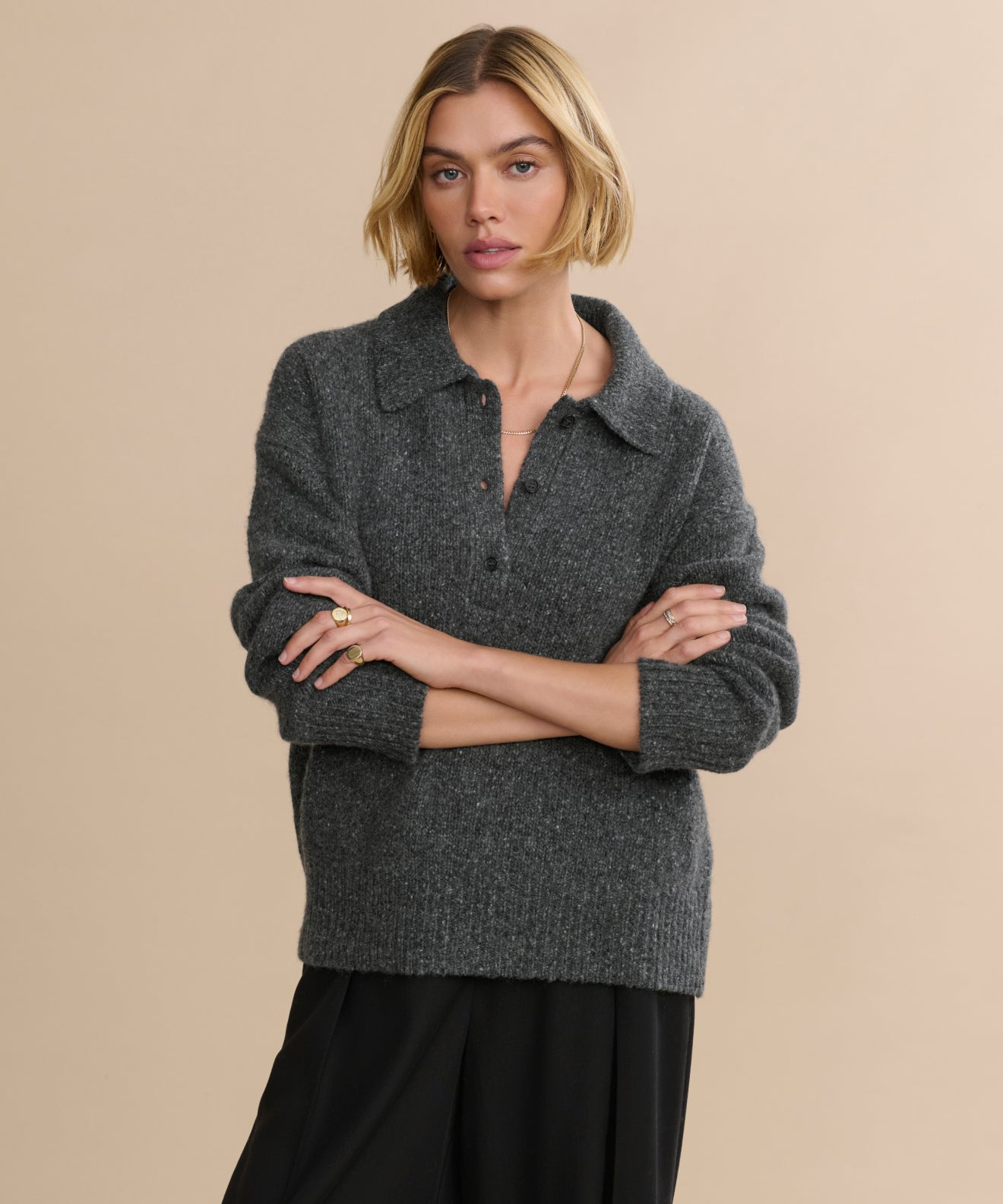 A woman with short blonde hair, wearing the Cashmere Burke Polo in dark grey and black pants, stands with arms crossed against a plain beige background.