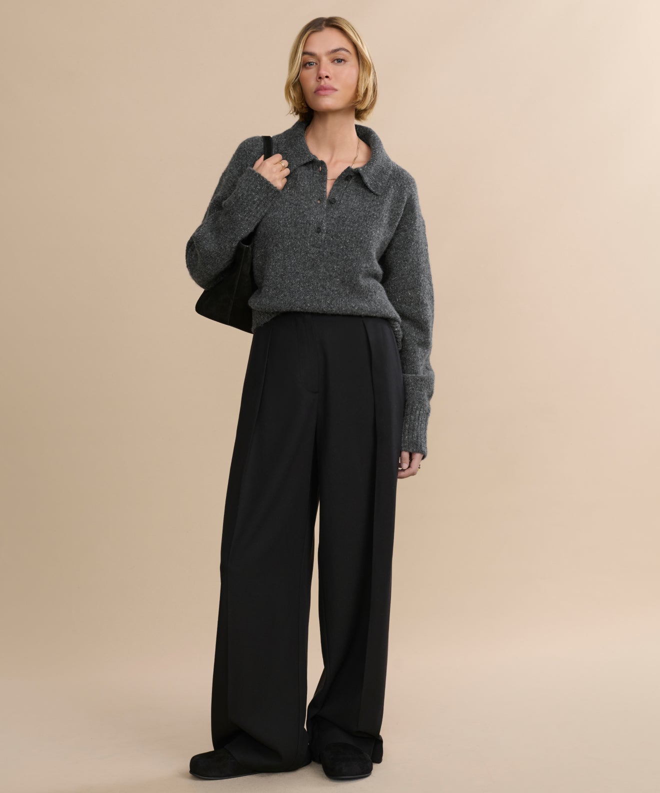 A woman with short blonde hair and a neutral expression stands against a beige background wearing the Cashmere Burke Polo, loose black trousers, black shoes, and carrying a black bag over her shoulder.
