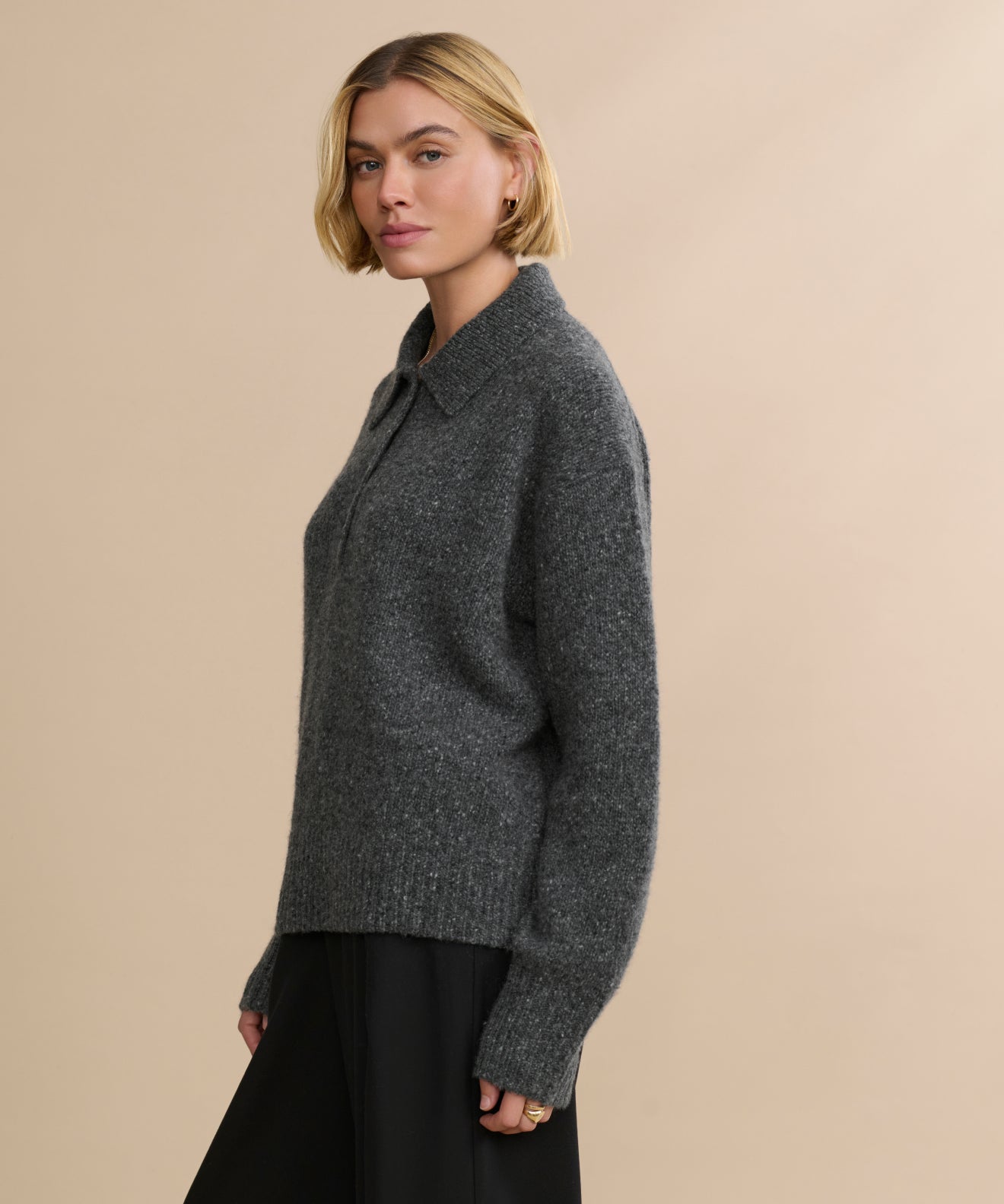 A woman with short blonde hair wears an oversized, dark gray Cashmere Burke Polo sweater with a collar and long sleeves, paired with black pants. She stands against a plain beige background, looking slightly to the side.