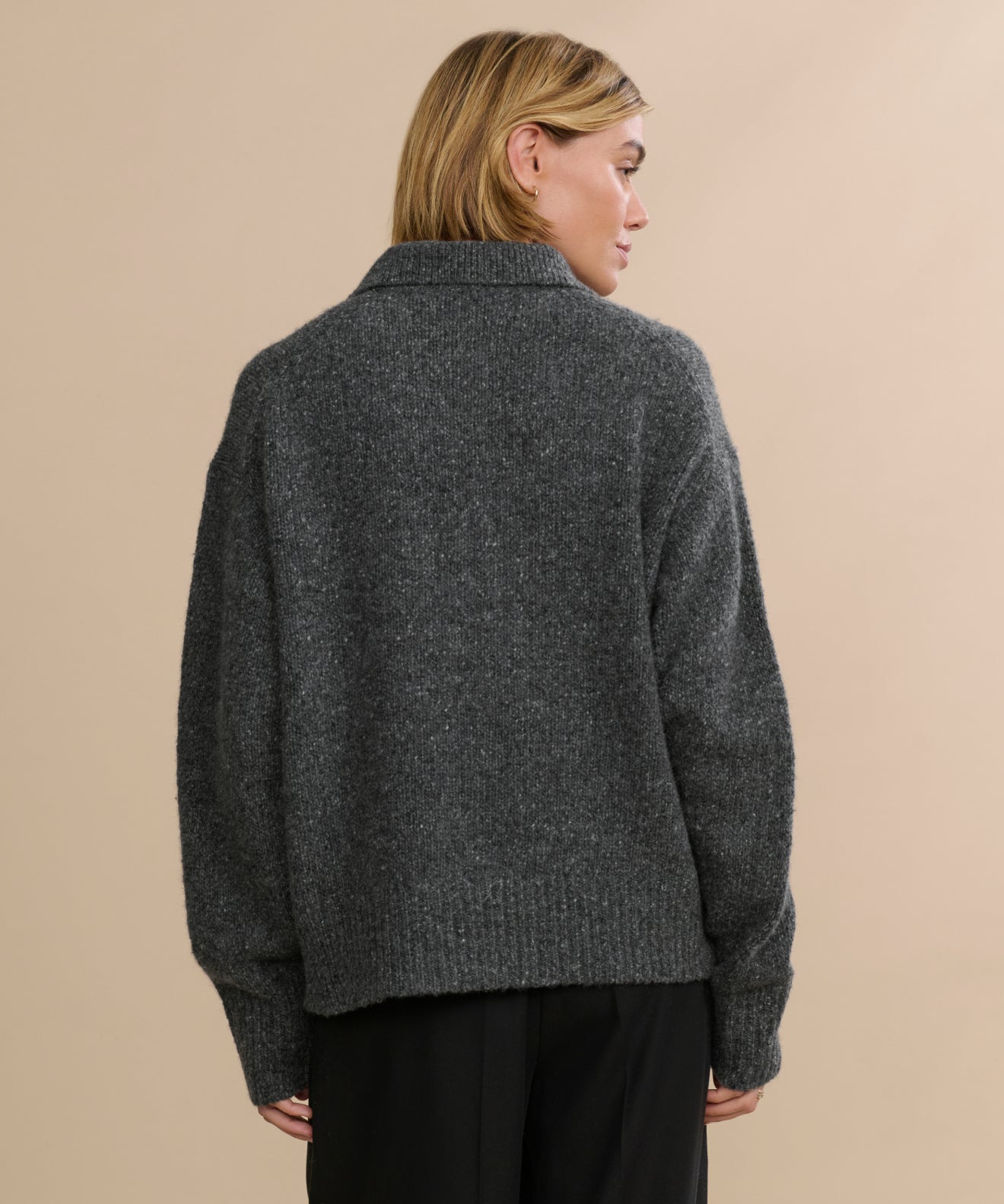 A person with short blonde hair, seen from behind, is wearing a Cashmere Burke Polo in dark gray with black pants against a plain, light beige background.