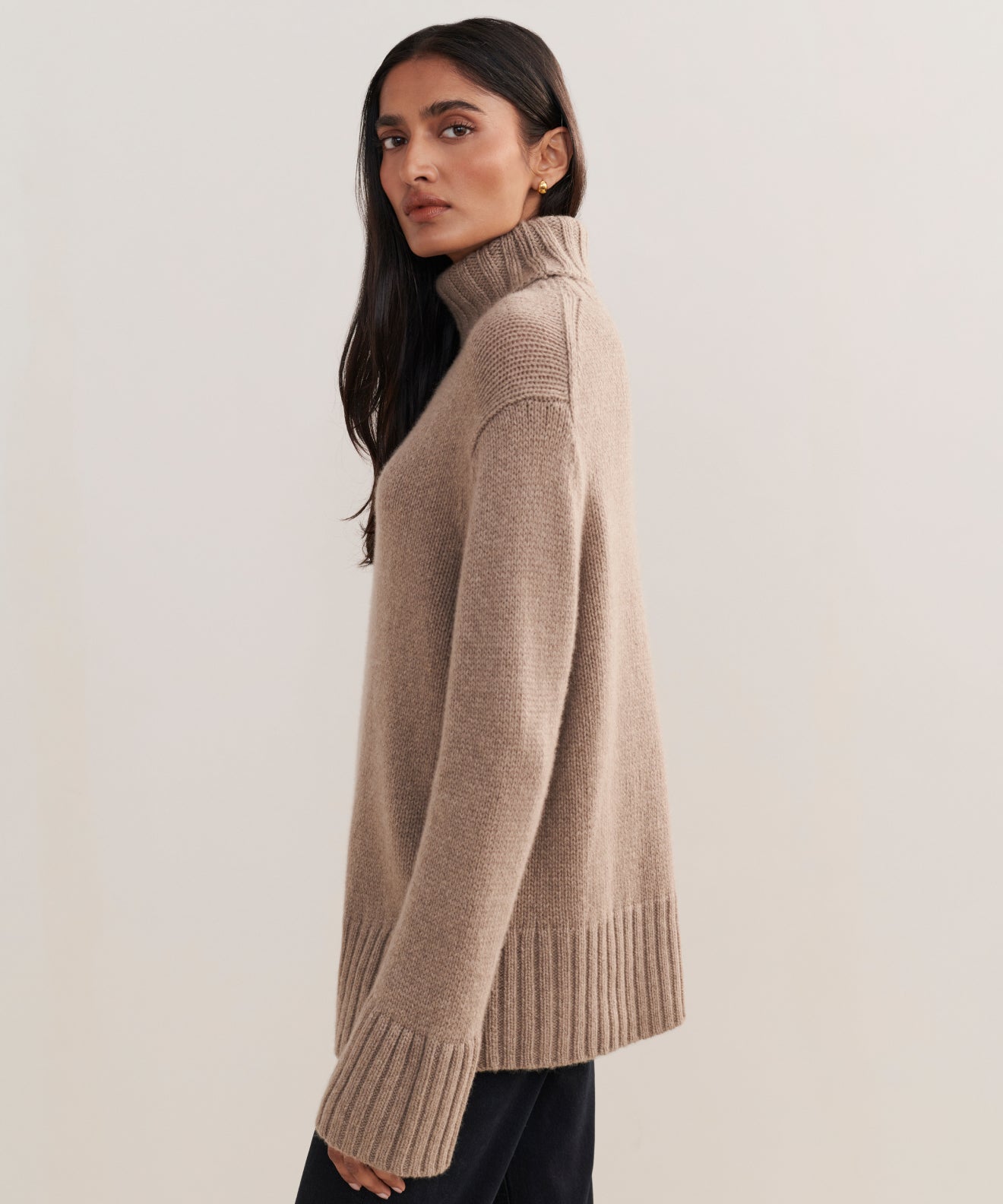 A woman with long dark hair models the Cashmere Jet Turtleneck, featuring ribbed cuffs and hem, as she stands sideways and looks at the camera against a plain light background.