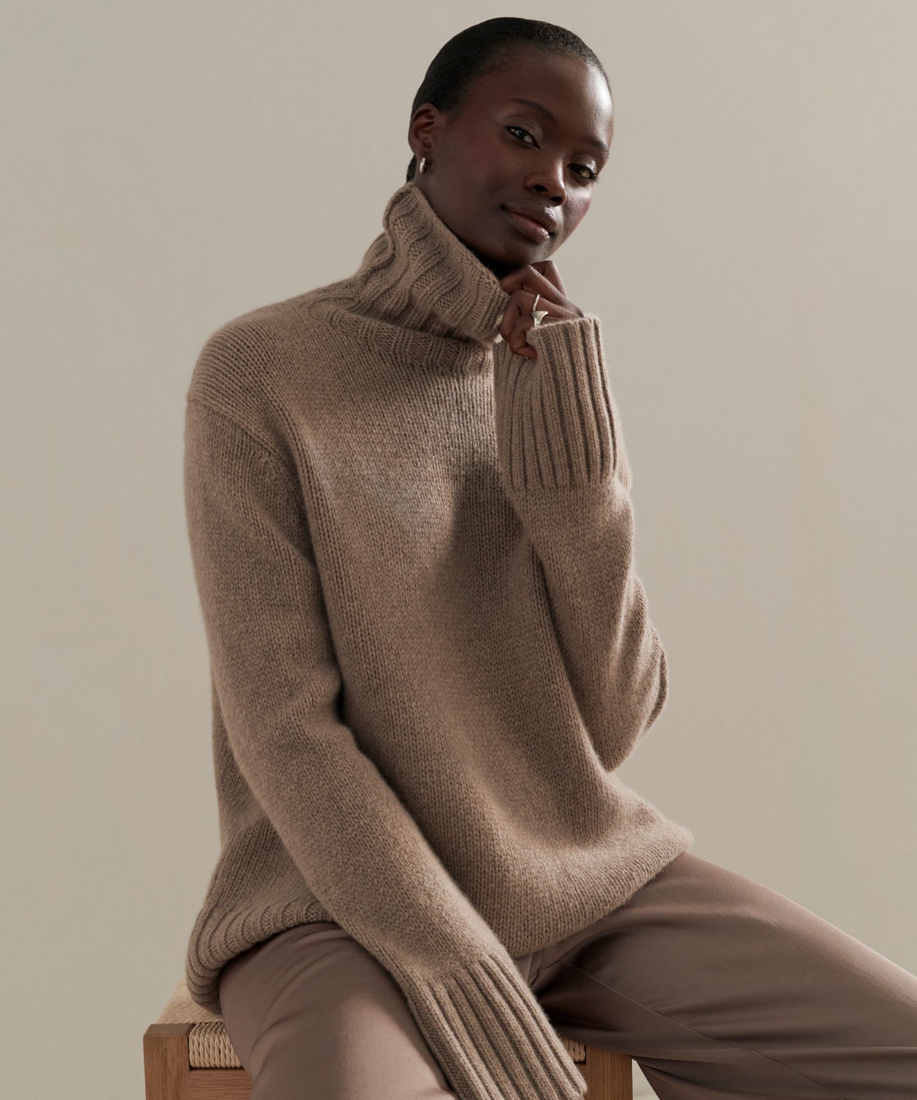 Seated on a wooden stool in a minimalist setting, a person wears the Cashmere Jet Turtleneck and matching pants, gently touching the collar of the cozy, oversized sweater.