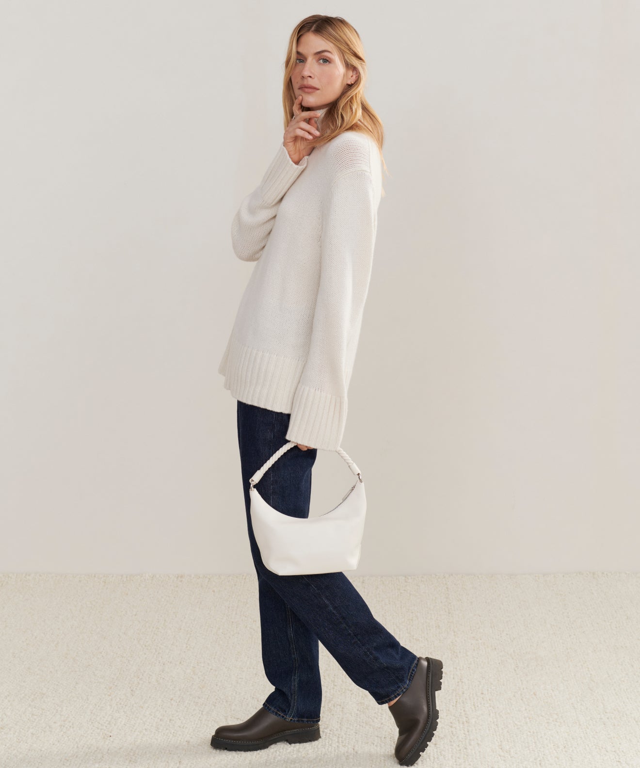 A woman stands sideways against a plain light background, wearing the Cashmere Jet Turtleneck with dark blue jeans and black boots. She holds a small white handbag and gazes forward, one hand near her chin.