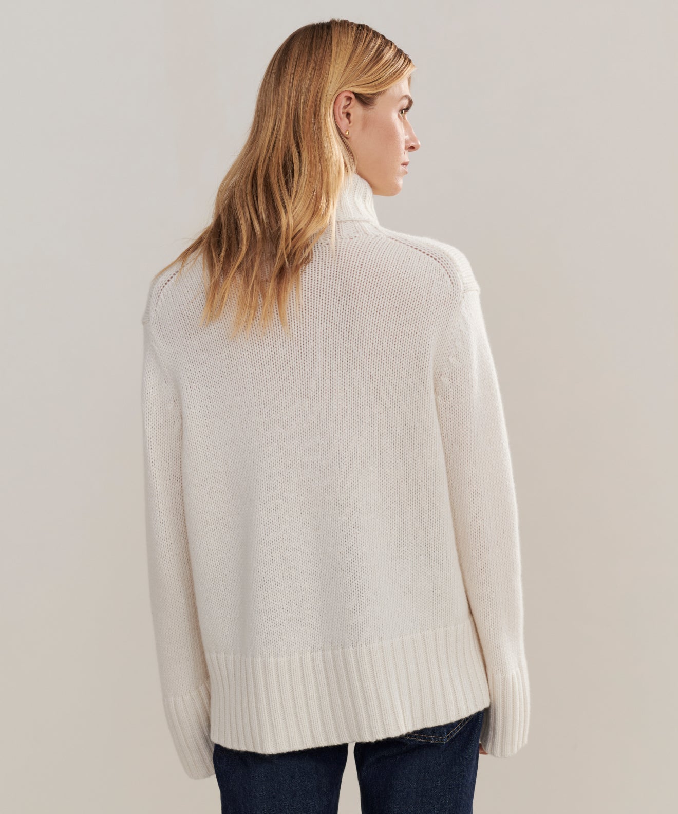 A person with long, blonde hair stands with their back to the camera against a plain light background, wearing the oversized Cashmere Jet Turtleneck and dark jeans.