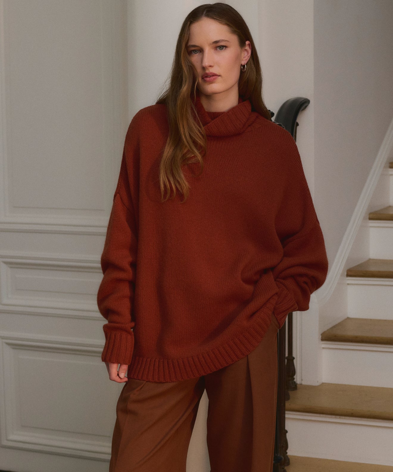 A woman with long brown hair stands indoors by a staircase, wearing the Cashmere Porter Turtleneck and wide-legged brown pants. She looks calmly at the camera, one hand in her pocket, effortlessly stylish.
