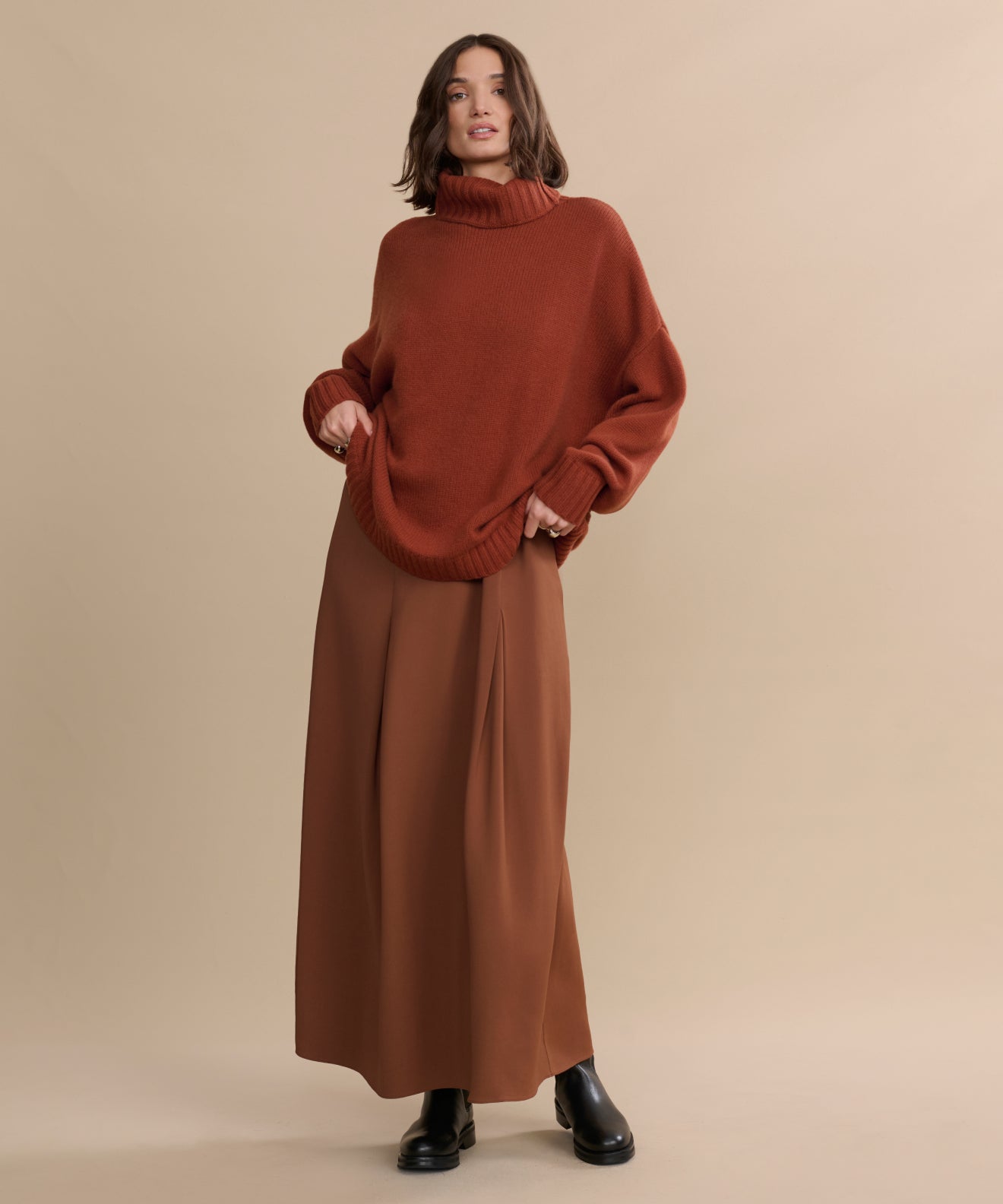 A person stands against a beige background, wearing the oversized Cashmere Porter Turtleneck with wide-legged brown pants and black boots, posing in a relaxed and confident manner.