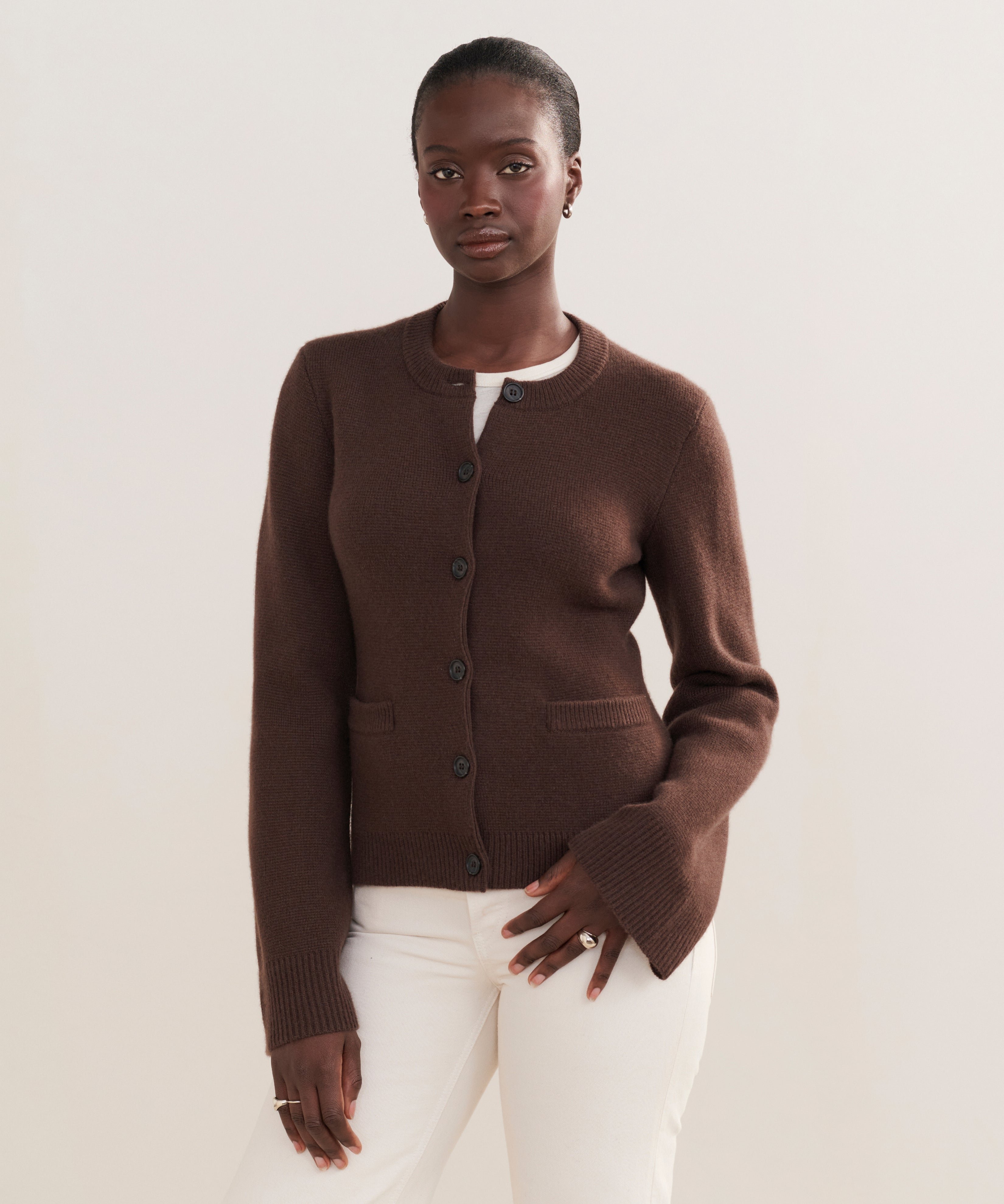 A woman wears the Cooper Cardigan, a dark brown button-up with a structured silhouette, over a white top and pants against a light background, illustrating its versatile styling for any occasion.