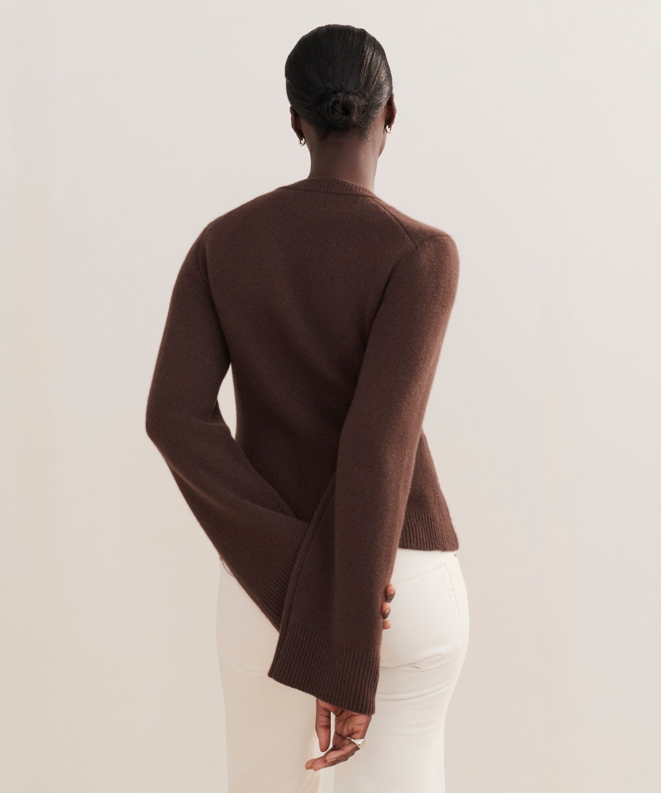 A person stands facing away, wearing the Cooper Cardigan in brown with a structured fit and off-white pants. Their hair is in a neat bun, one hand behind their back, against a plain, light-colored background.