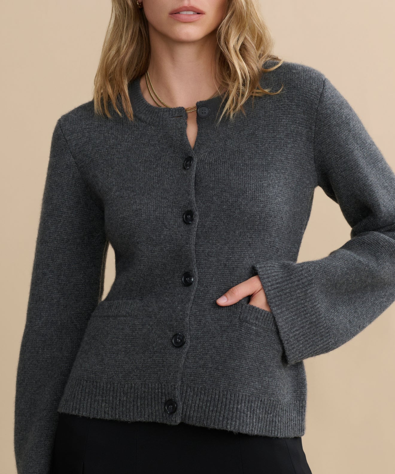 A woman models the Cooper Cardigan, a dark grey button-up with flared sleeves and two front pockets, standing against a beige background. Her left hand is in the pocket, and her face is partially out of frame.