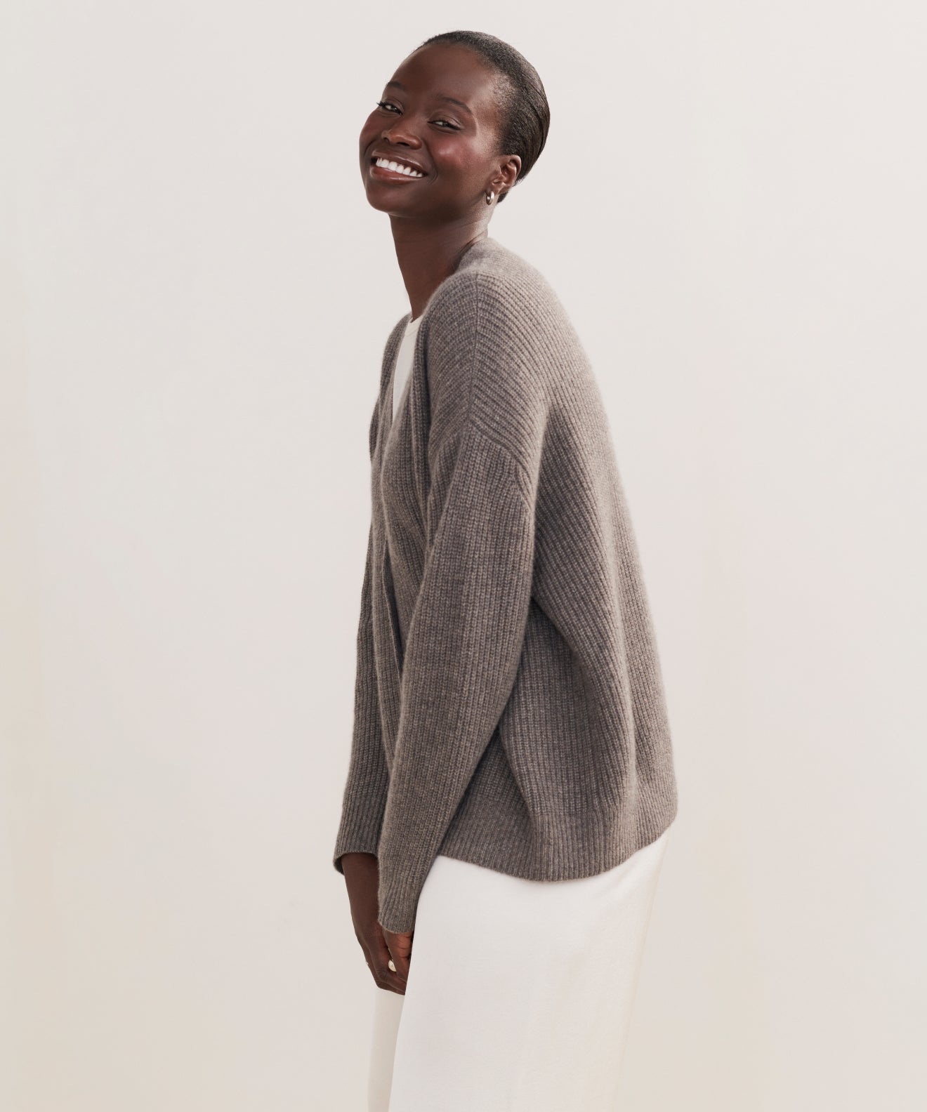A woman with short hair smiles while standing and tilting slightly forward. She wears the Cropped Cashmere Cocoon Cardigan paired with a white skirt against a plain light background.