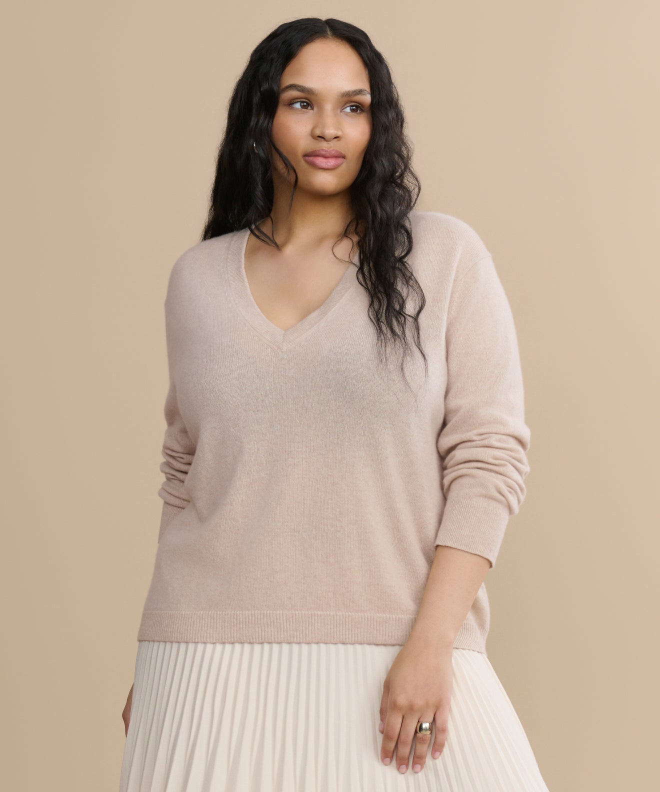 A woman with long wavy hair wears the Flynn Cashmere Sweater in light beige along with a pleated cream skirt, standing against a plain beige background and gazing to the side with a relaxed expression.