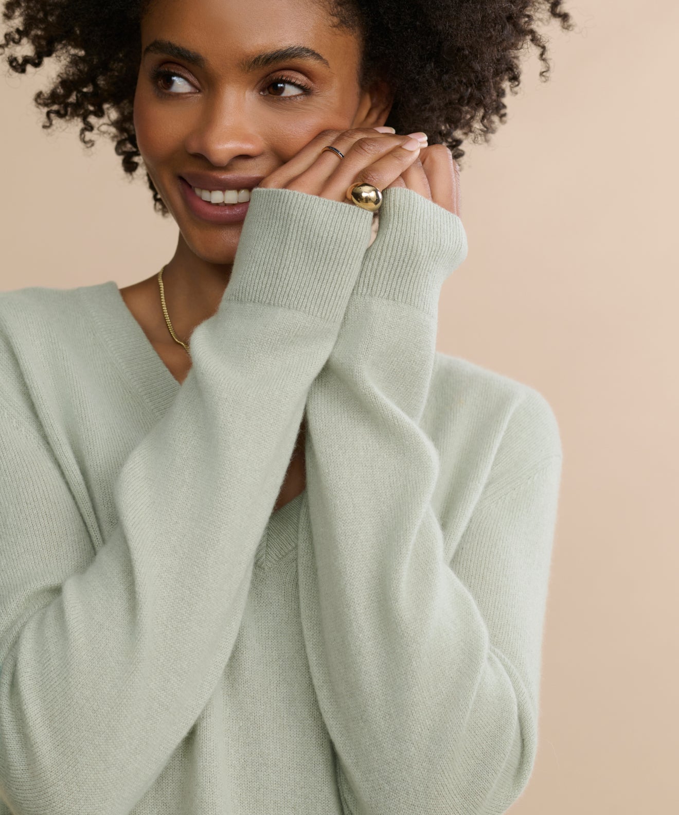 A woman with curly hair smiles gently, wearing the Flynn Cashmere Sweater in light sage green with a relaxed fit and long sleeves, paired with a gold ring on her finger, posed against a beige background.