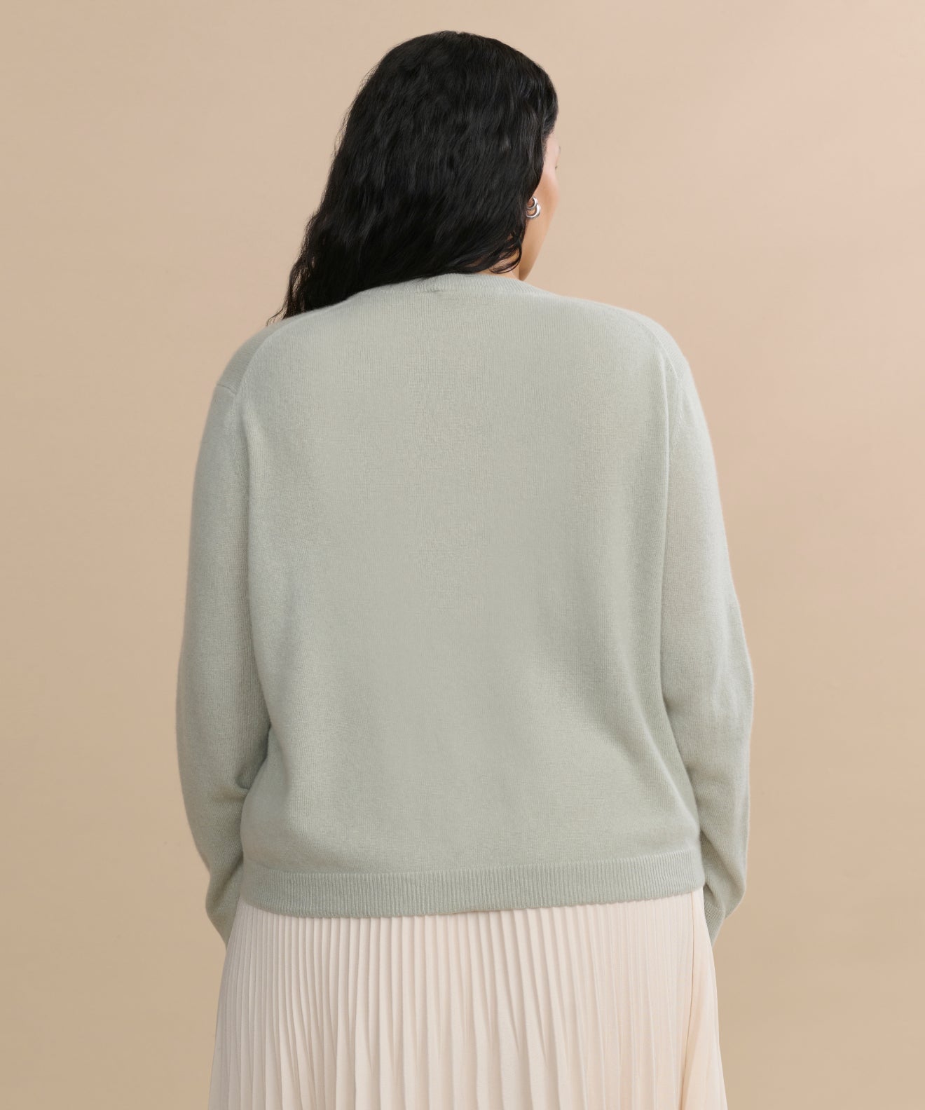 A person with long, wavy dark hair is seen from behind wearing the Flynn Cashmere Sweater in light grayish-green with a cream pleated skirt, set against a plain beige background.