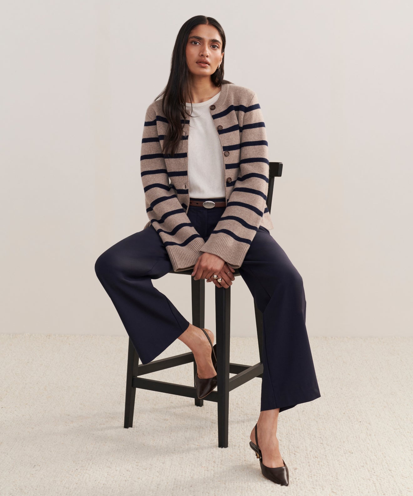 A woman with long dark hair sits on a tall black chair, wearing the Cooper Cardigan in taupe/navy stripe over a white top, navy wide-leg pants, and brown shoes. She faces forward with a neutral expression.