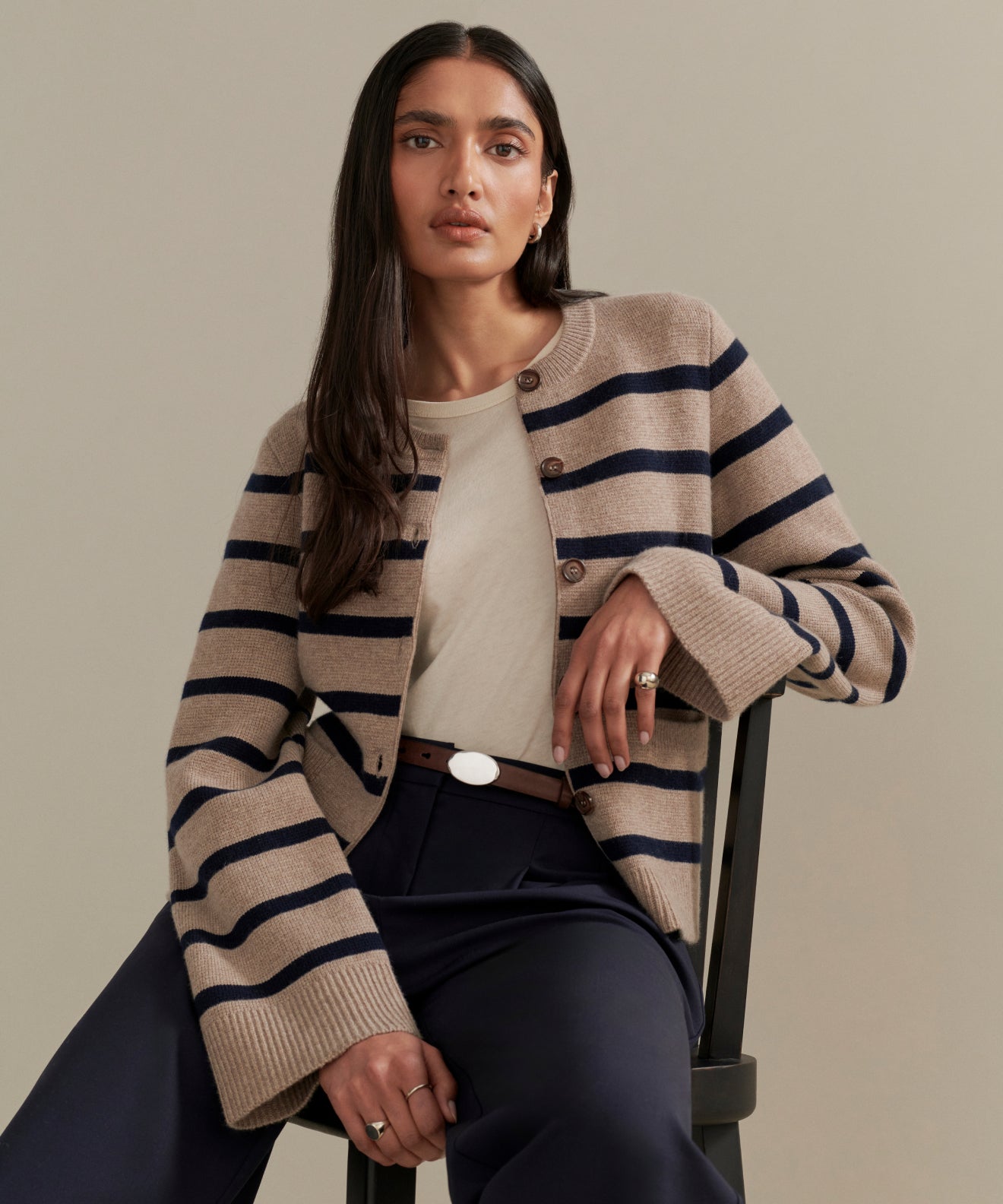 A woman with long, dark hair sits on a chair, calmly modeling the Cooper Cardigan in Taupe/Navy Stripe over a cream top and navy pants, highlighting its structured silhouette.