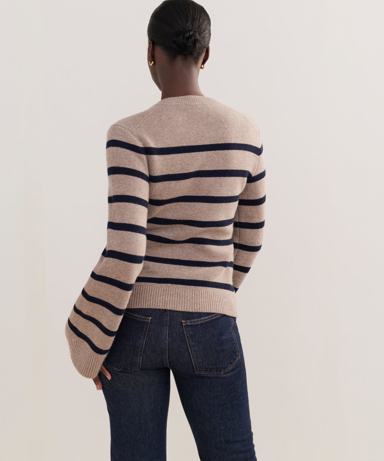 A person with hair in a low bun faces away, wearing the Cooper Cardigan in Taupe/Navy Stripe, featuring wide horizontal stripes, a structured silhouette, and slightly flared sleeves, paired with dark blue jeans.